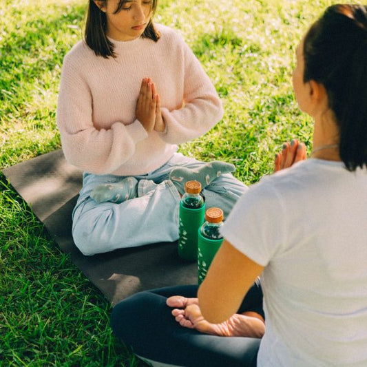 zwei frauen meditieren im freien auf yogamatten mit grünen wasserflaschen aus glas